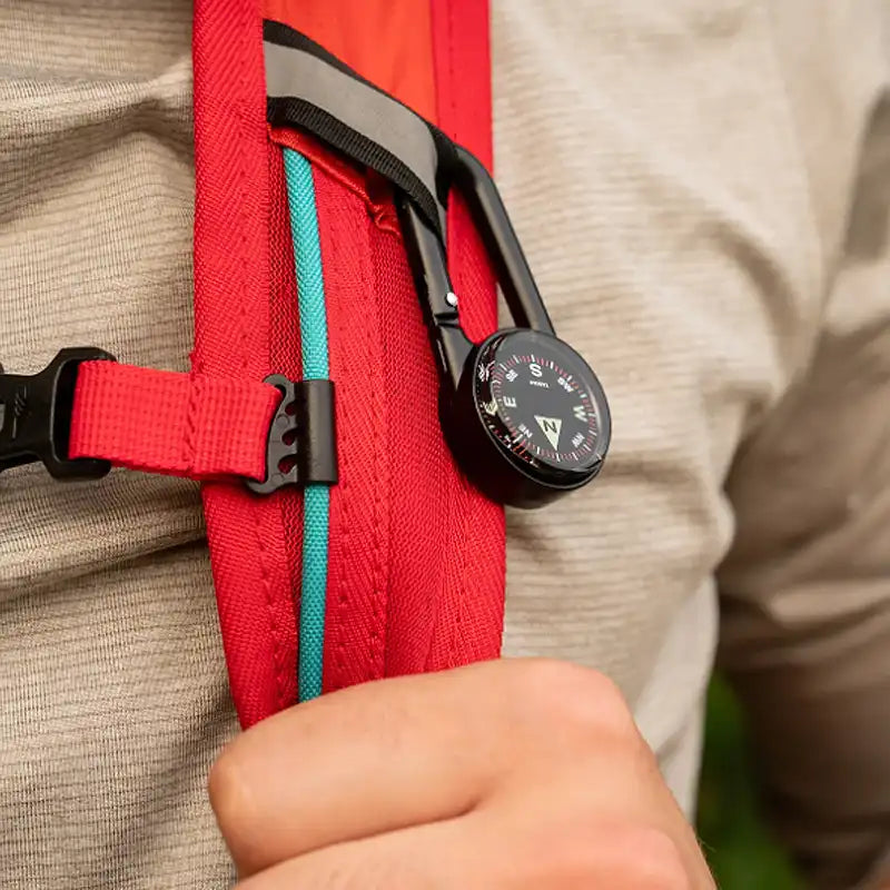 A close-up of a red backpack strap with a black carabiner holding a black compass.