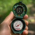 A green folding pocket compass with an integrated thermometer, held in a person's hand against a blurred green outdoor background.